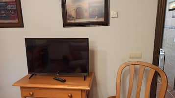 Un televisor sobre un soporte de madera, un control remoto, una silla y una pared con cuadros enmarcados.