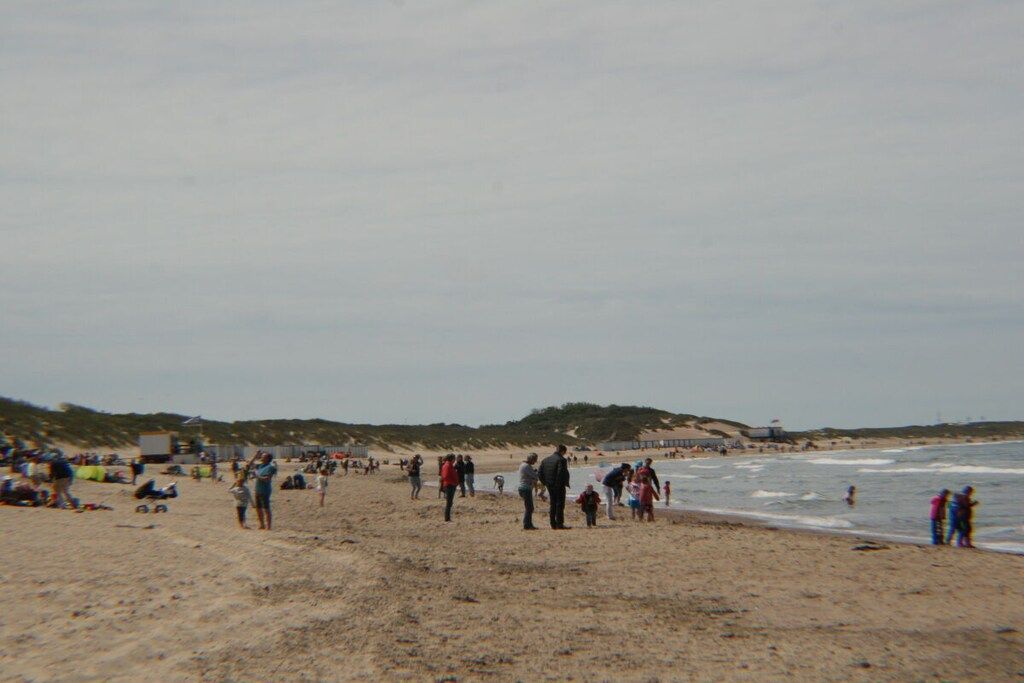 A beach with people walking and standing, a lighthouse in the distance, and dunes in the background.