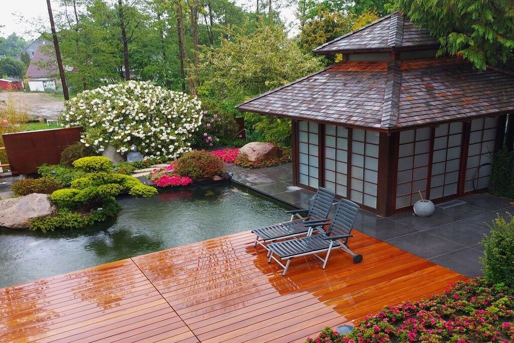 A traditional Japanese-style building with a tiled roof and wooden lattice walls, adjacent to a wooden deck with a small pond and lush greenery.