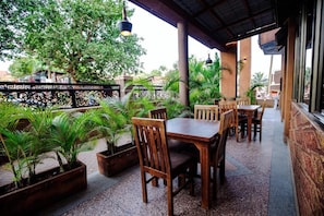 Un patio al aire libre con mesas y sillas de madera, rodeado de plantas en macetas y una valla decorativa.