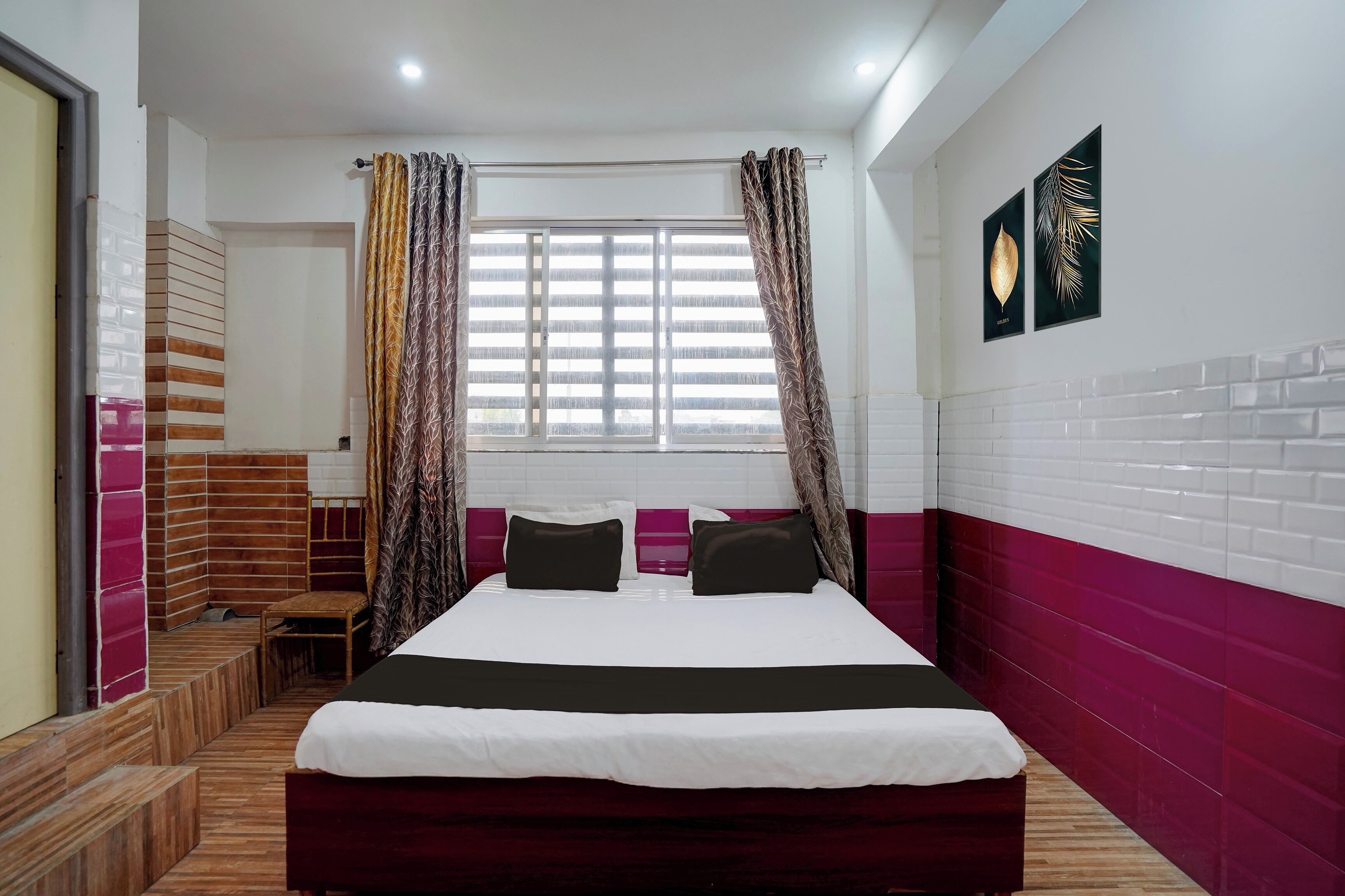 A hotel room with a bed, a window with curtains, a chair, and a wall with two framed pictures.