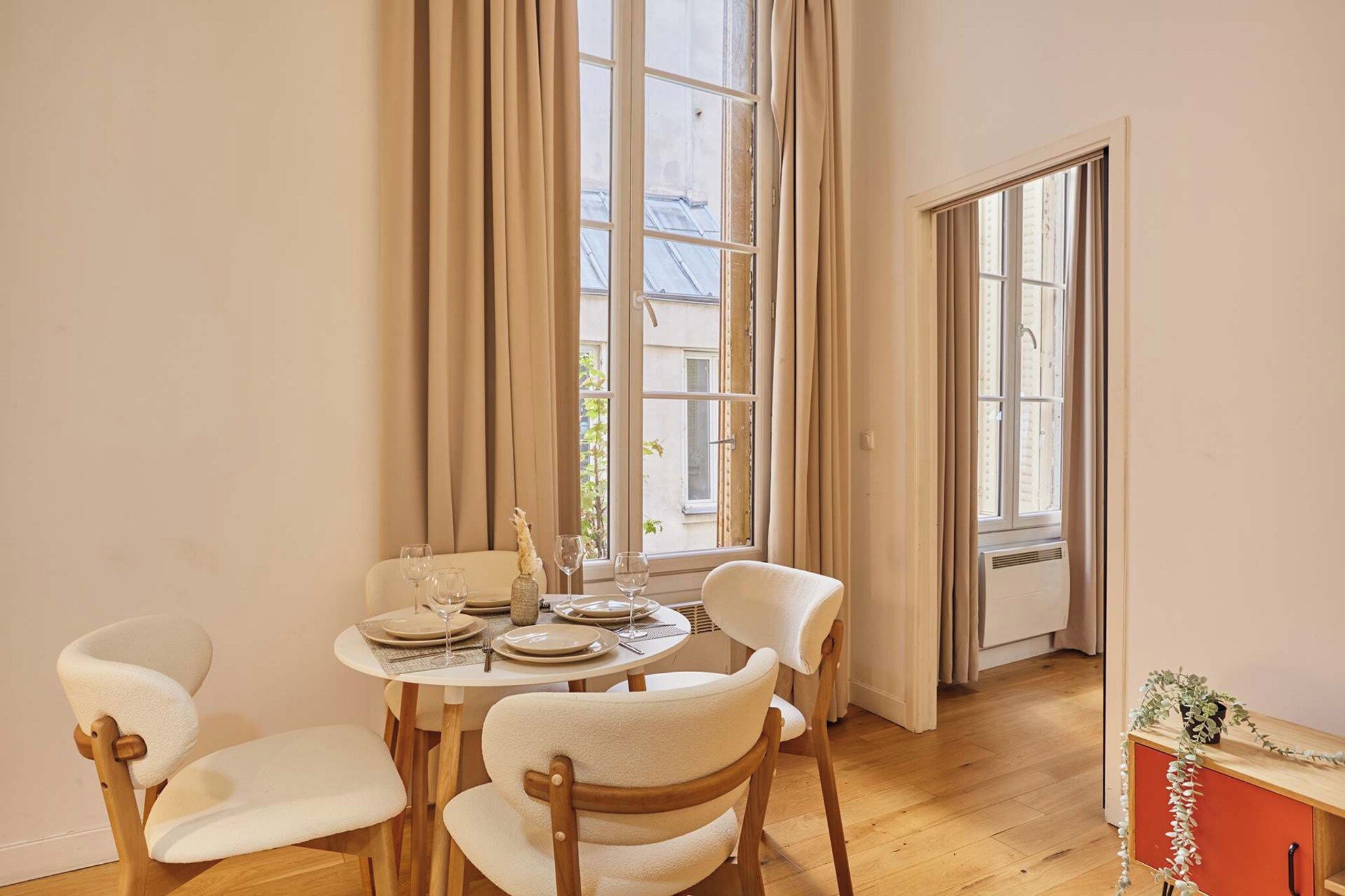 Un coin repas avec une table ronde dressée pour quatre personnes, entourée de chaises, et de grandes fenêtres agrémentées de rideaux.