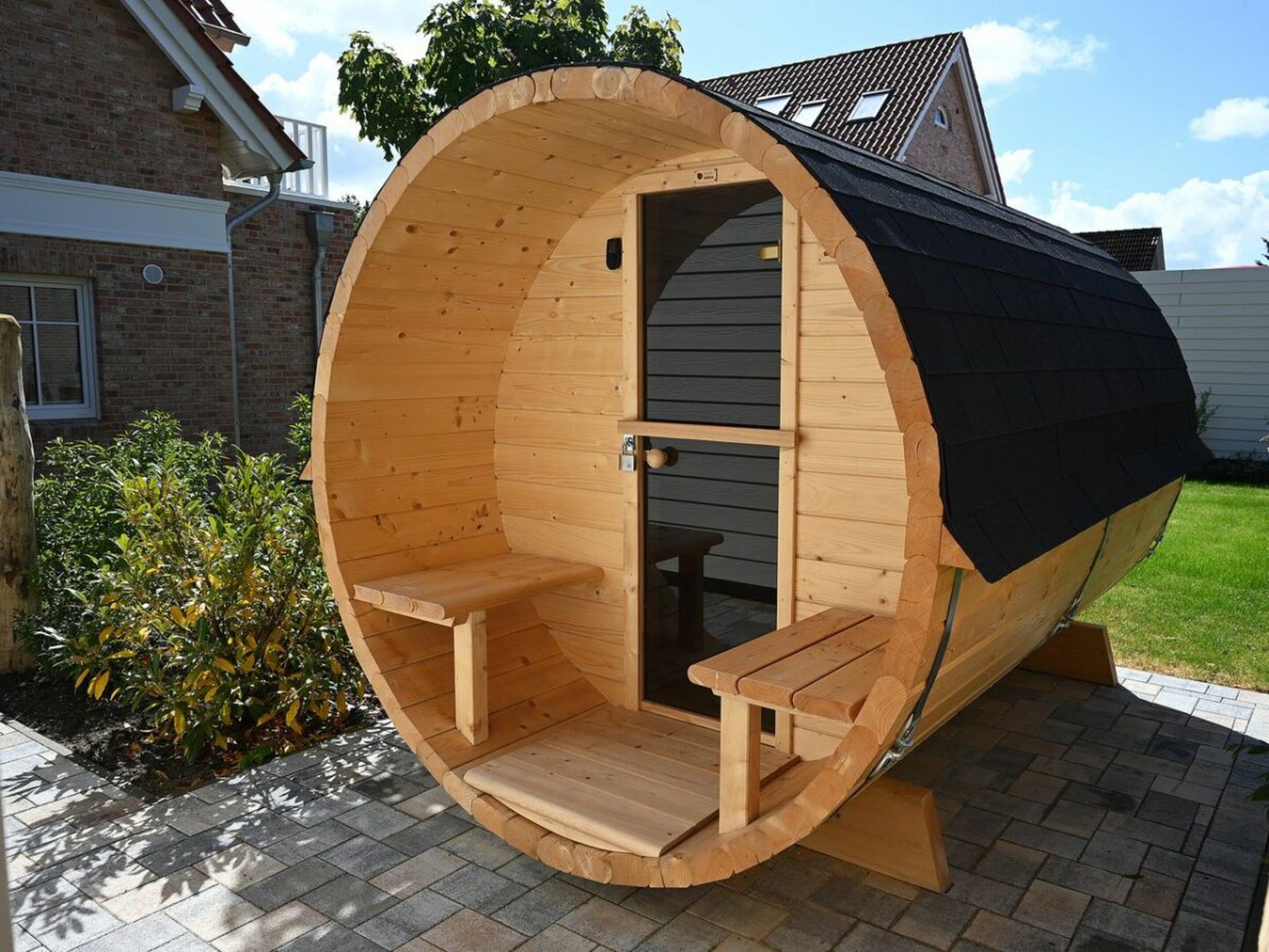A wooden sauna with two benches and a door, situated on a paved area with a house in the background.