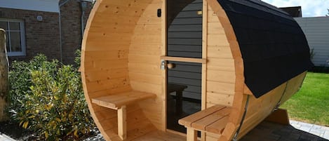 A wooden sauna with two benches and a door, situated on a paved area with a house in the background.