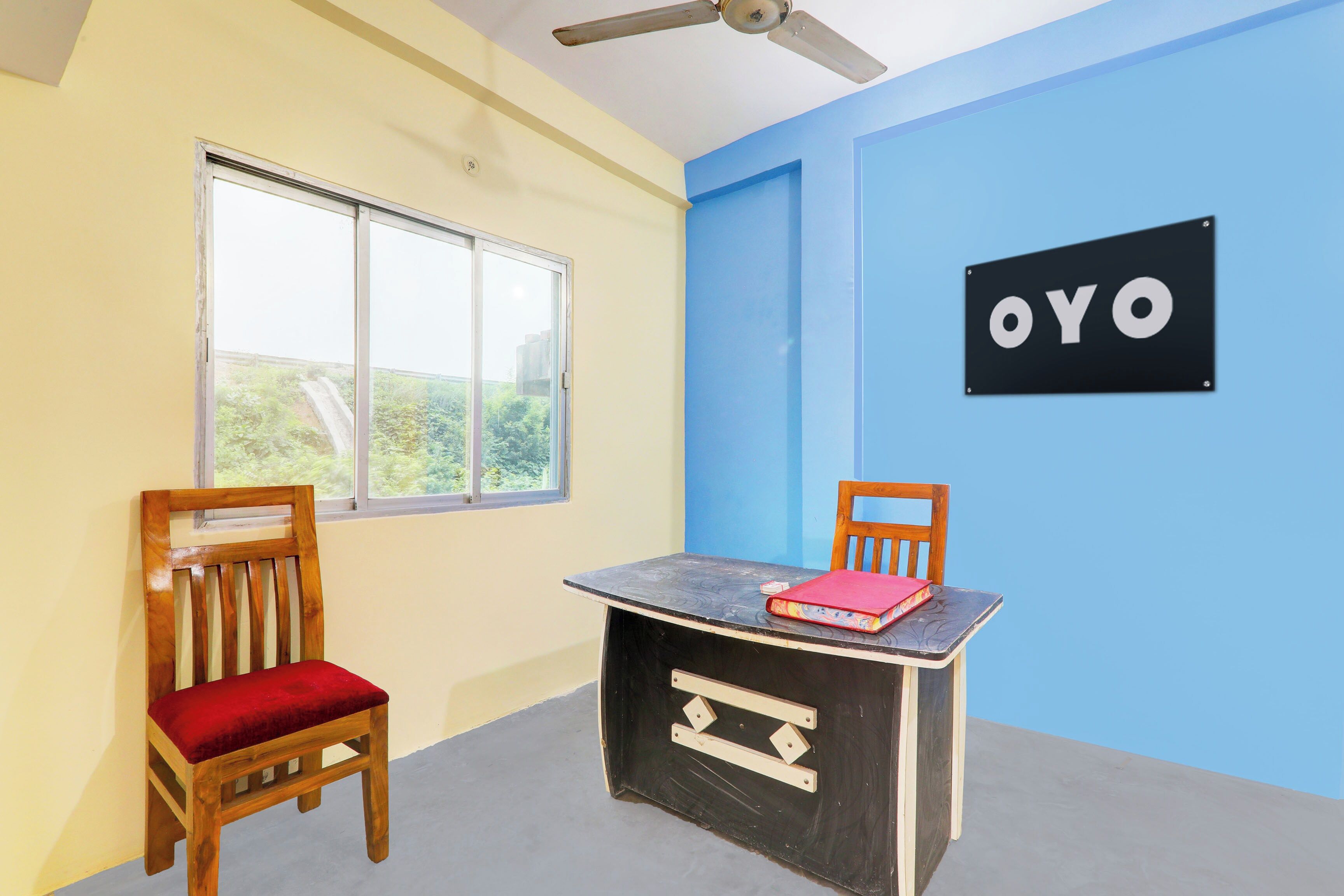 A room with a single window, a ceiling fan, a table with a red book, and a wall sign that reads 'OYO'.