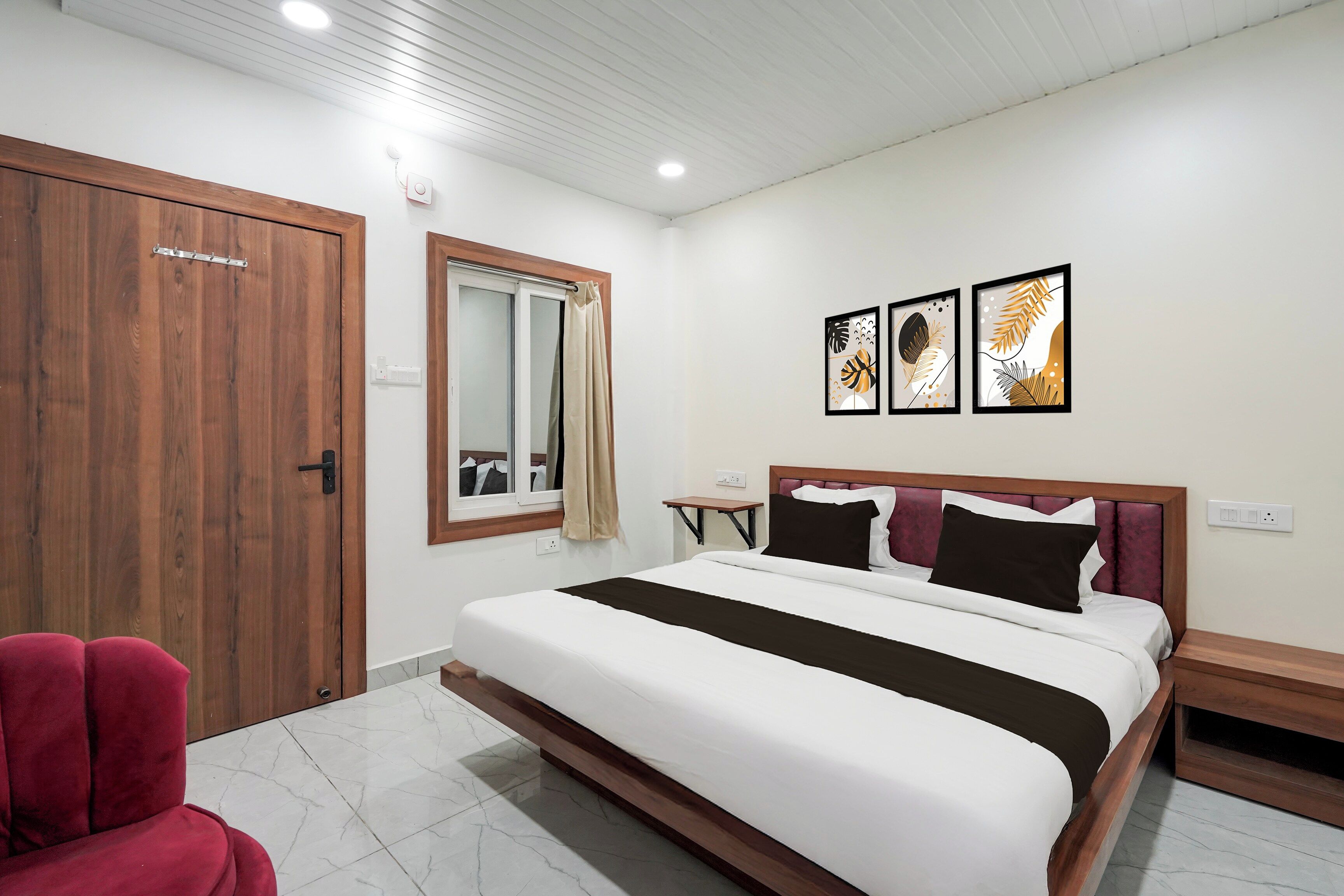 A hotel room with a bed, a wooden wardrobe, a red chair, a small table, and artwork on the wall.