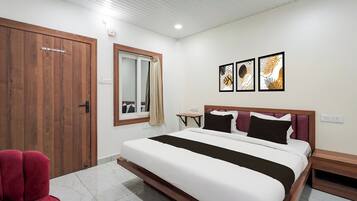 A hotel room with a bed, a wooden wardrobe, a red chair, a small table, and artwork on the wall.