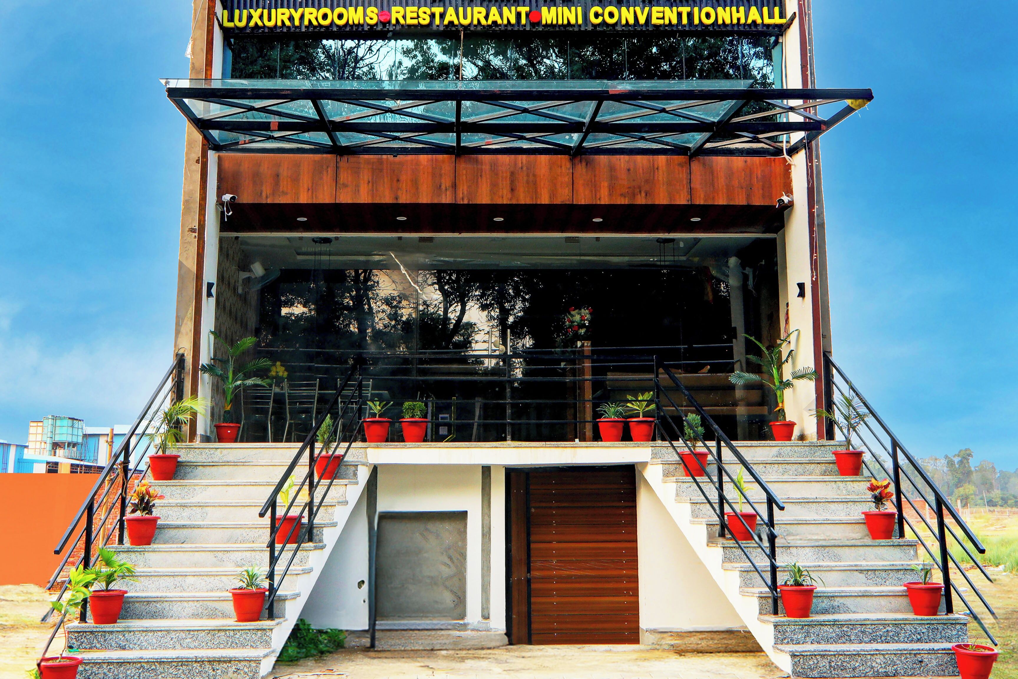 A building with a sign reading 'LUXURYROOMS RESTAURANT MINI CONVENTIONHALL'. It features a staircase with red planters, a wooden door, and a glass facade.