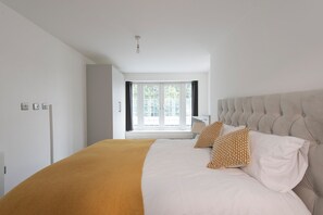 Een slaapkamer met een groot bed, een raam met gordijnen, een plafondlamp en een deur naar een andere kamer.
