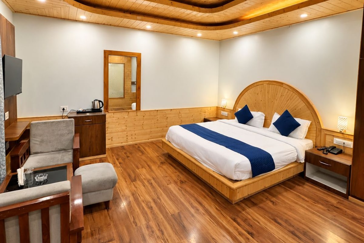 A bedroom with a wooden ceiling, a large bed with blue bedding, a bedside table, a chair, and a small table with a lamp.