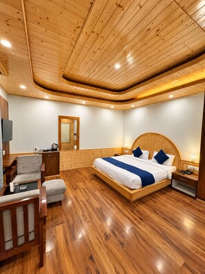 A bedroom with a wooden ceiling, a large bed with blue bedding, a bedside table, a chair, and a small table with a lamp. - Hotel Old Manali Retreat (Manali)