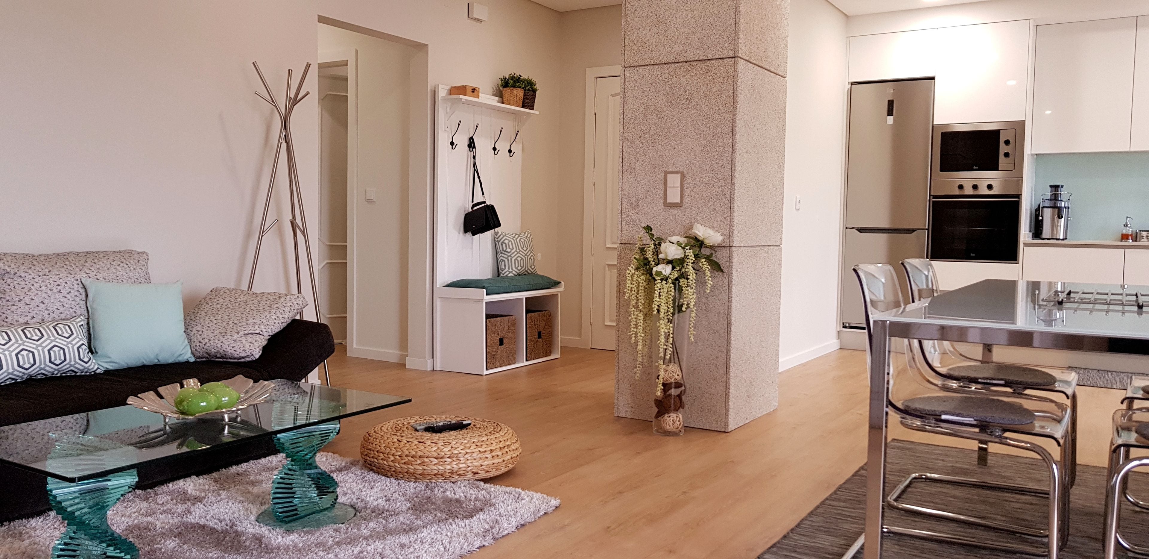 A modern living room with a glass coffee table, a sofa with patterned cushions, a woven pouf, and a decorative vase with flowers.