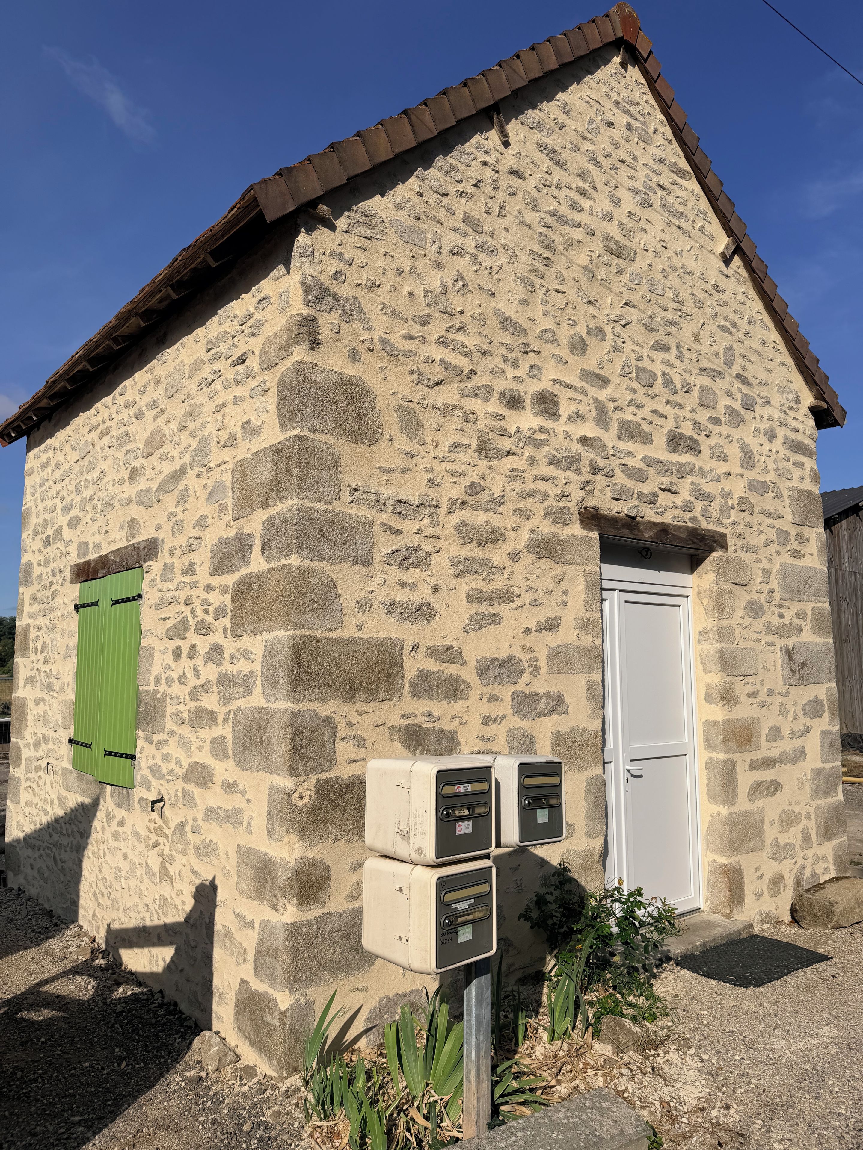 Un bâtiment en pierre avec un volet vert, une porte blanche et un coffret de raccordement contenant des compteurs.