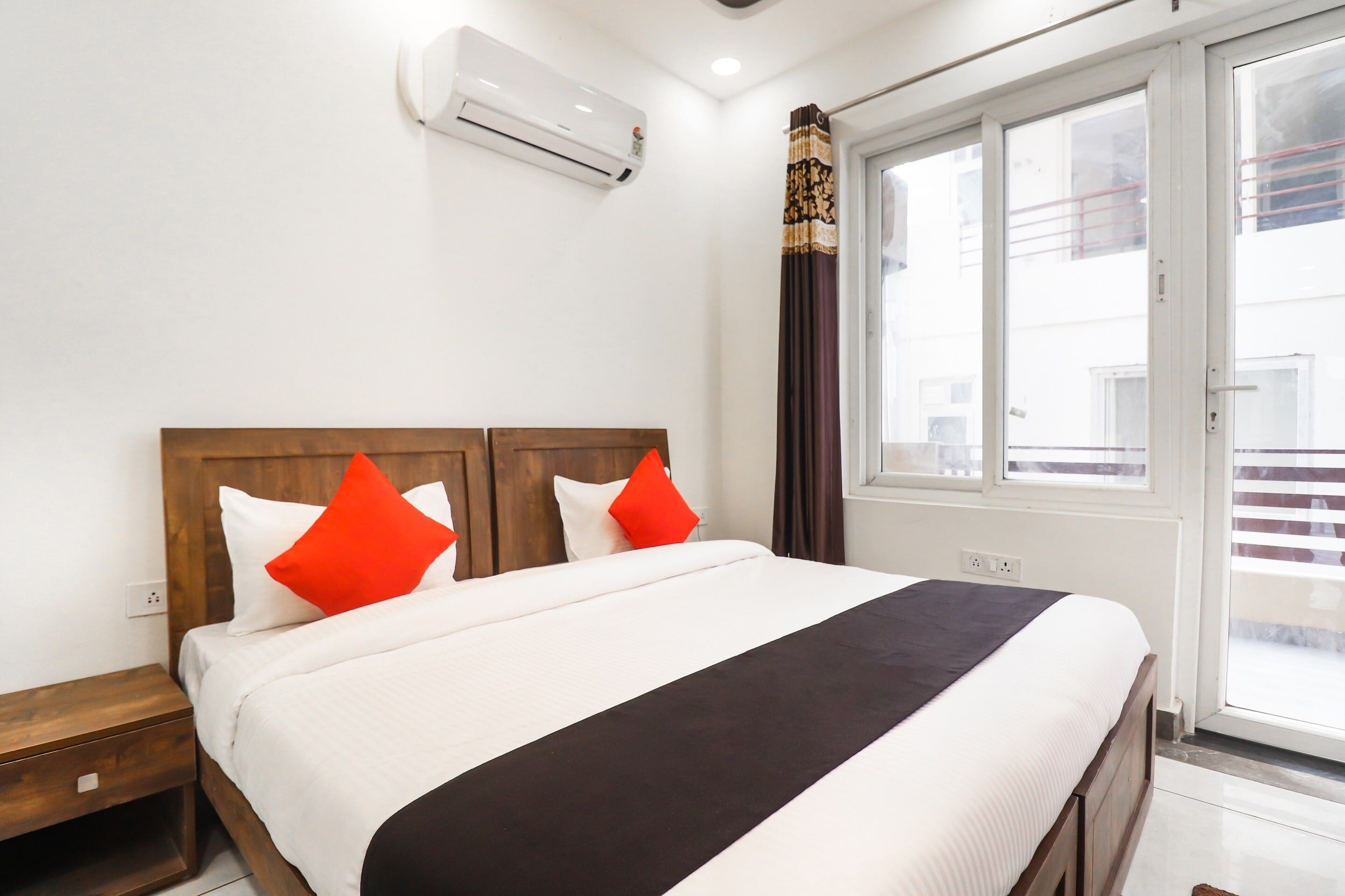 A hotel room with a bed, a wooden headboard, a window with curtains, and an air conditioning unit on the wall.
