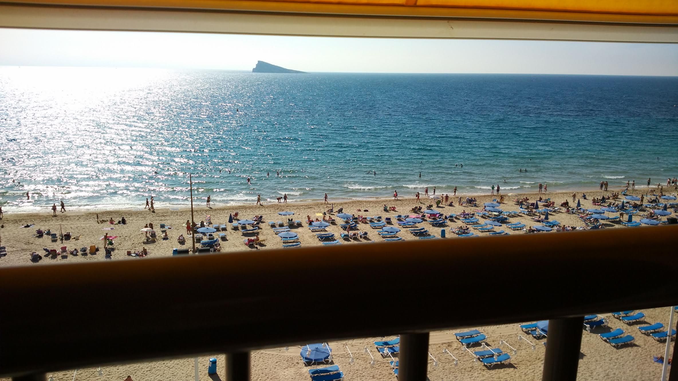 A beach with numerous sun loungers and umbrellas, a clear view of the ocean, and a distant island.
