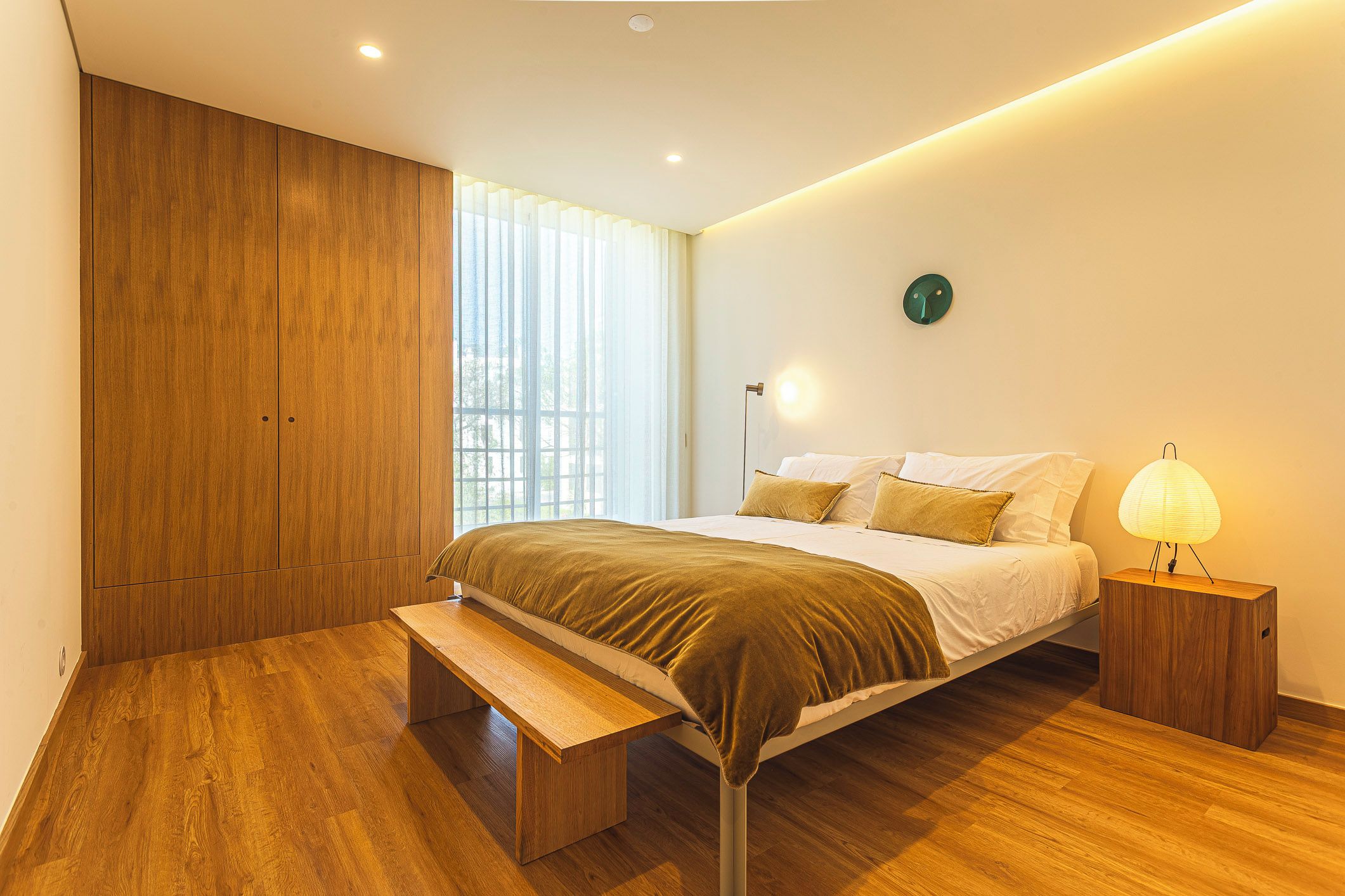Un dormitorio con cama, mesita de noche, banco, armario de madera y ventana con cortinas.