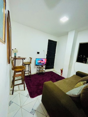 Un espacio habitable compacto con una cama, un televisor sobre un mueble, una mesita con sillas y un jarrón decorativo.