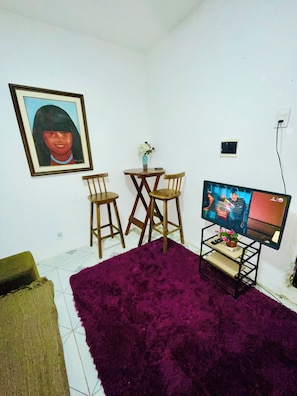 Una habitación con una televisión sobre un mueble bajo, dos sillas de madera, una mesita con un florero, un cuadro en la pared y una alfombra morada.