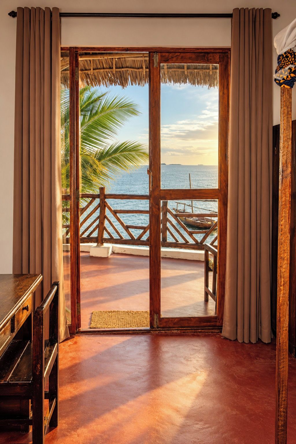 Quarto com porta de madeira que dá para uma varanda com vista para o mar e palmeiras.