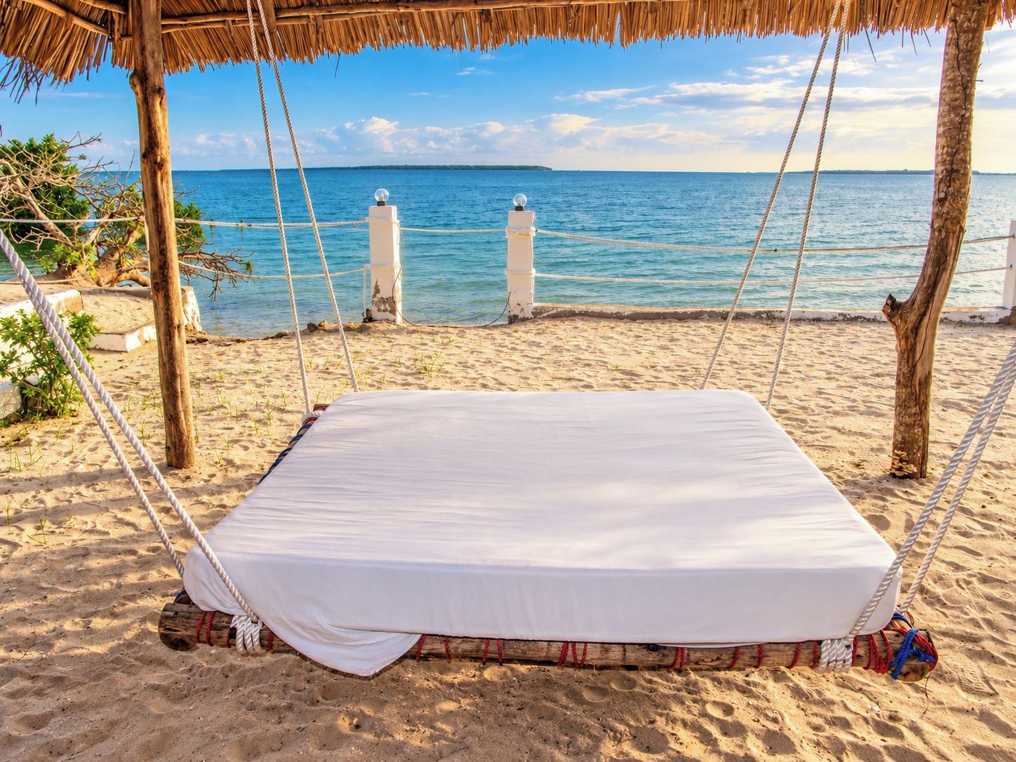 A beach hammock bed with a white mattress, ropes, and a thatched roof structure.