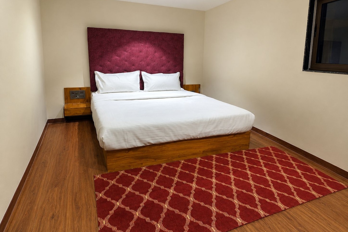 A hotel room with a large bed, a red patterned rug, wooden flooring, and a cream-colored wall.