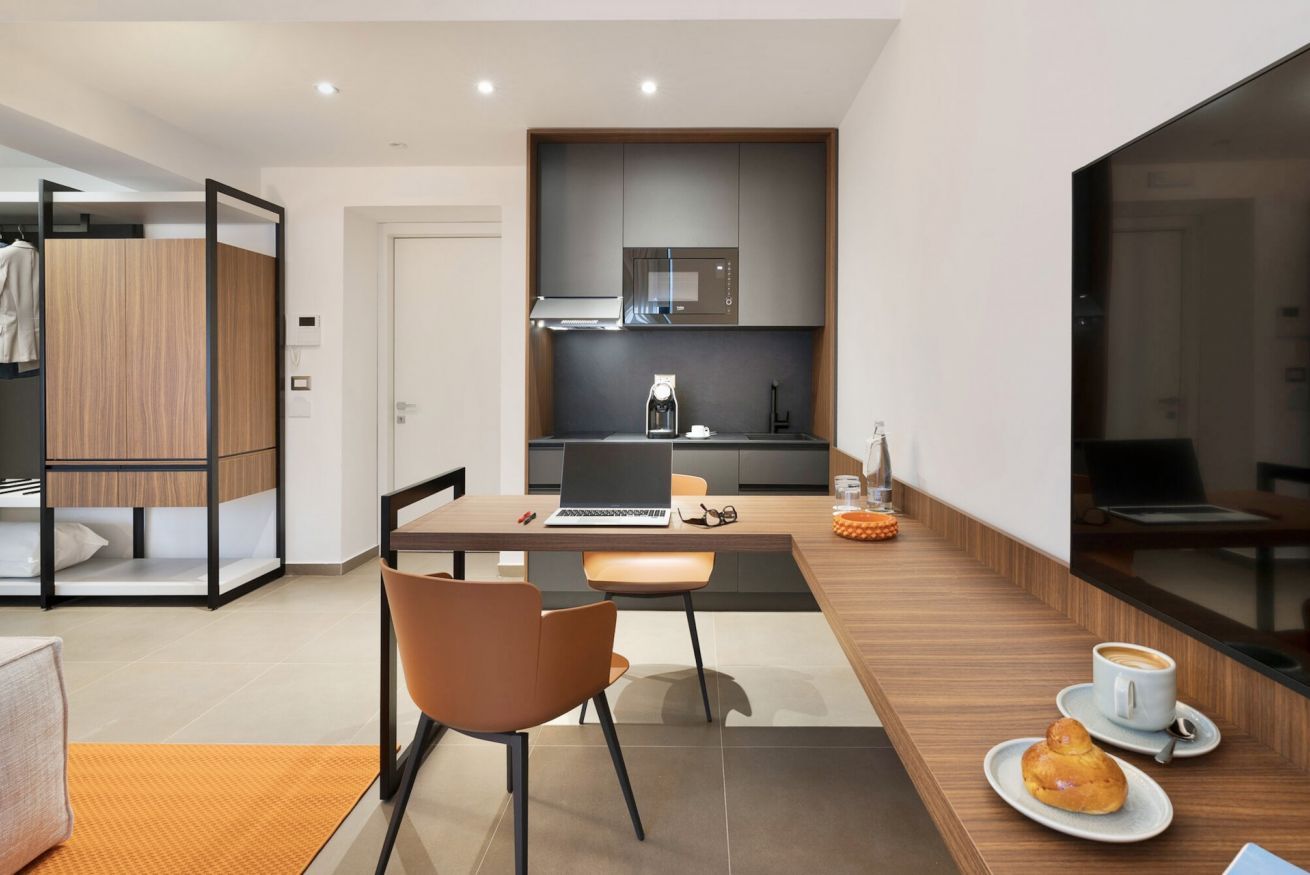 A modern hotel room with a dining area, kitchenette, and a large window.