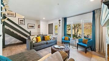 A spacious living room with a grey sofa, blue armchairs, and a coffee table. A staircase with a dark railing is visible on the left.