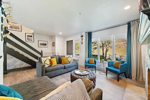 Ein geräumiges Wohnzimmer mit einem grauen Sofa, blauen Sesseln und einem Couchtisch. Eine Treppe mit dunkler Geländer ist links zu sehen.