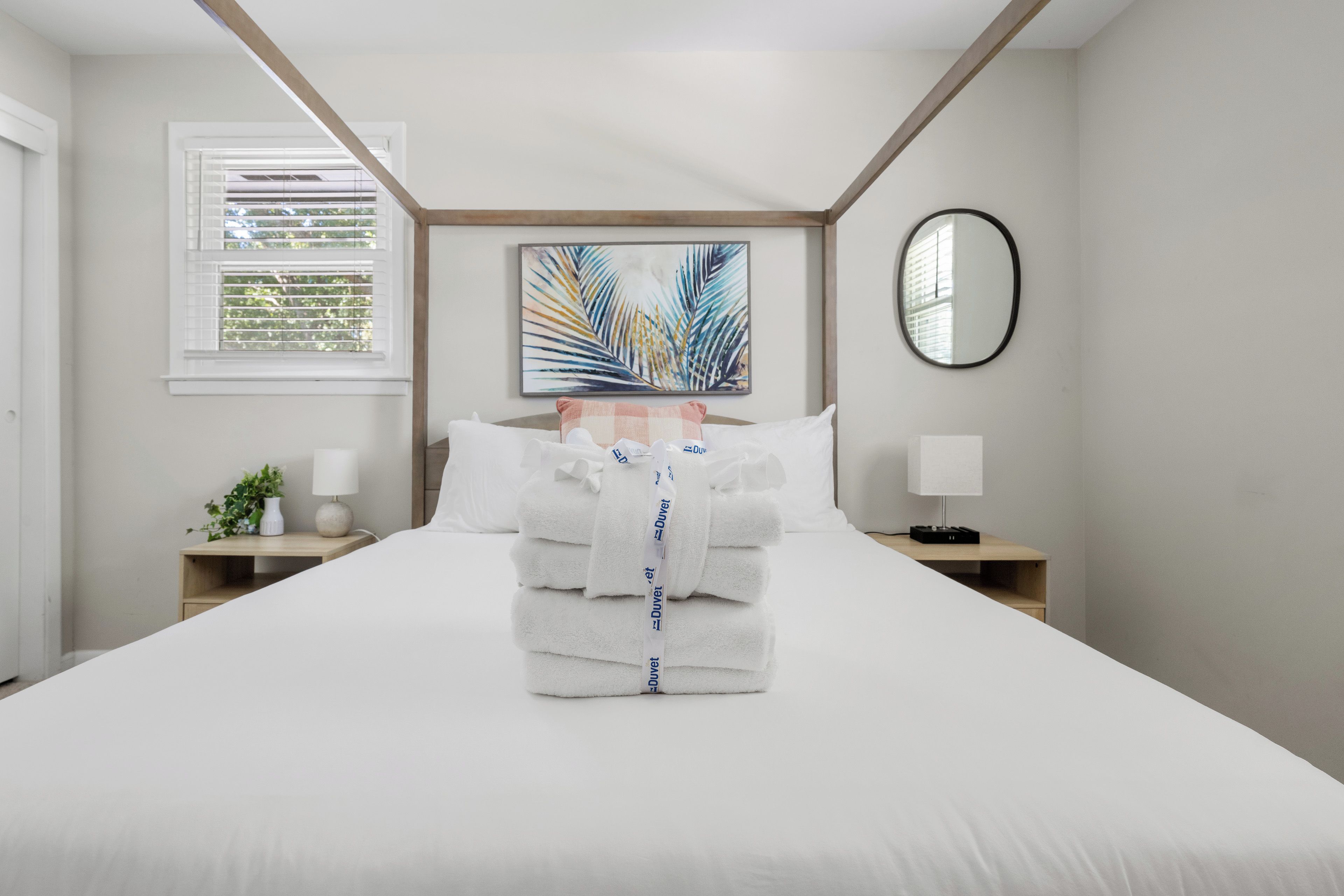 A neatly made bed with folded towels and a decorative palm leaf painting above it.