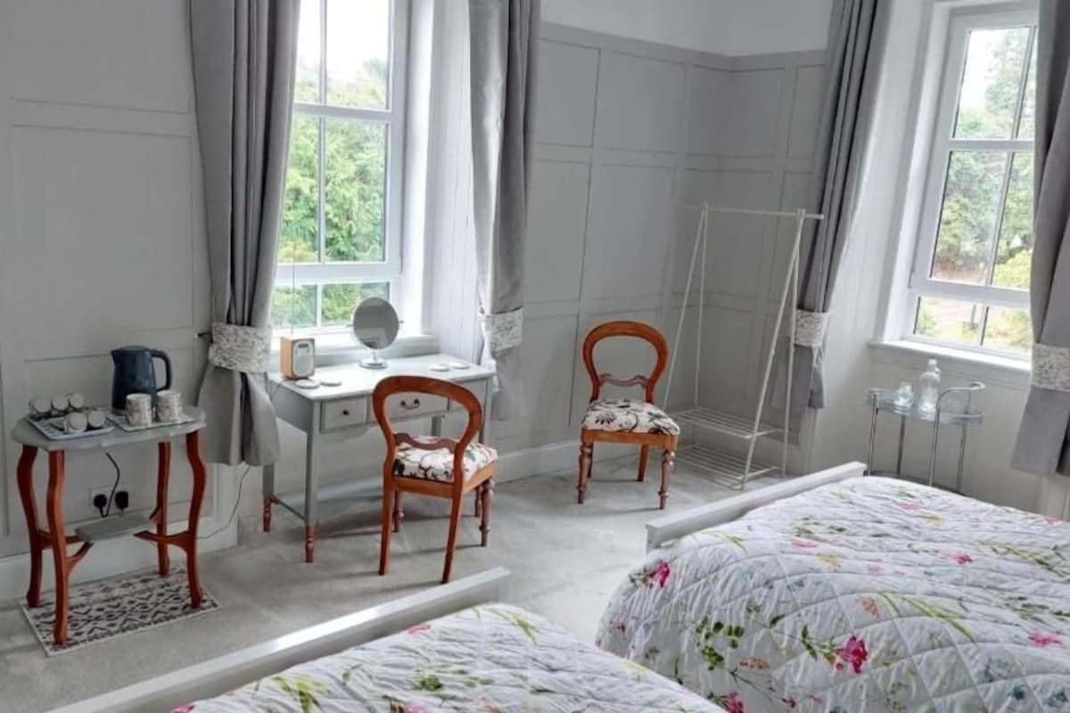 Une chambre à coucher avec un lit, un bureau avec une chaise, un miroir et une fenêtre avec des rideaux.