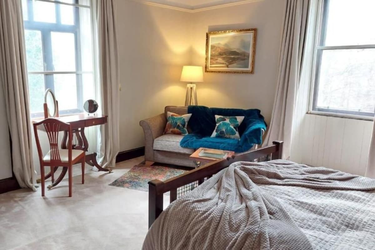 Une chambre d’hôtel avec un lit, un canapé, une table et une chaise. Il y a une fenêtre avec des rideaux, une lampe et un tableau au mur.