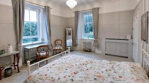Une chambre à coucher avec un grand lit, deux fenêtres, un radiateur, une chaise, une petite table et un miroir.