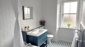 Une salle de bain avec un mur recouvert de carreaux blancs, un meuble-lavabo bleu, des toilettes blanches, une fenêtre avec une plante et un sol à motifs.