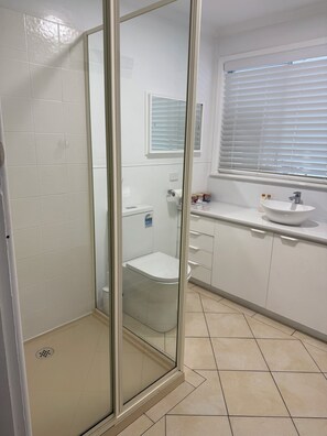Ett badrum med en dusch som är avskärmad med glasväggar, en toalett, ett handfat och ett fönster med persienner.