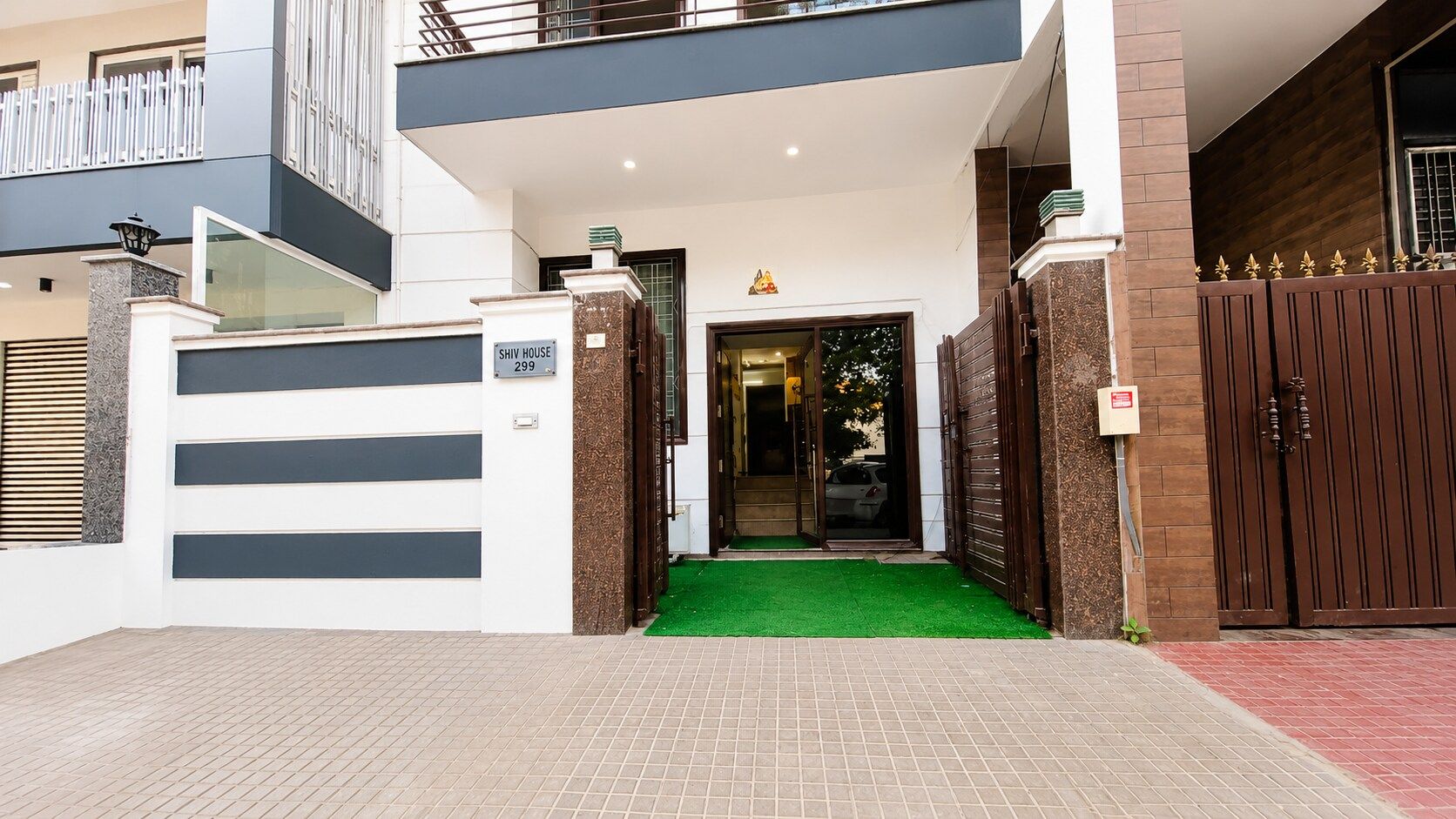 Une maison moderne avec une entrée sécurisée par un portail, un tapis vert menant à la porte et un balcon doté d’une balustrade.