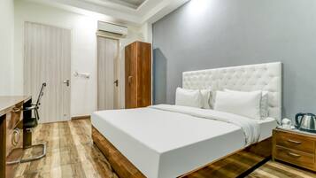 A hotel room with a large bed, wooden flooring, a wardrobe, a desk with a chair, and a bedside table with a kettle.