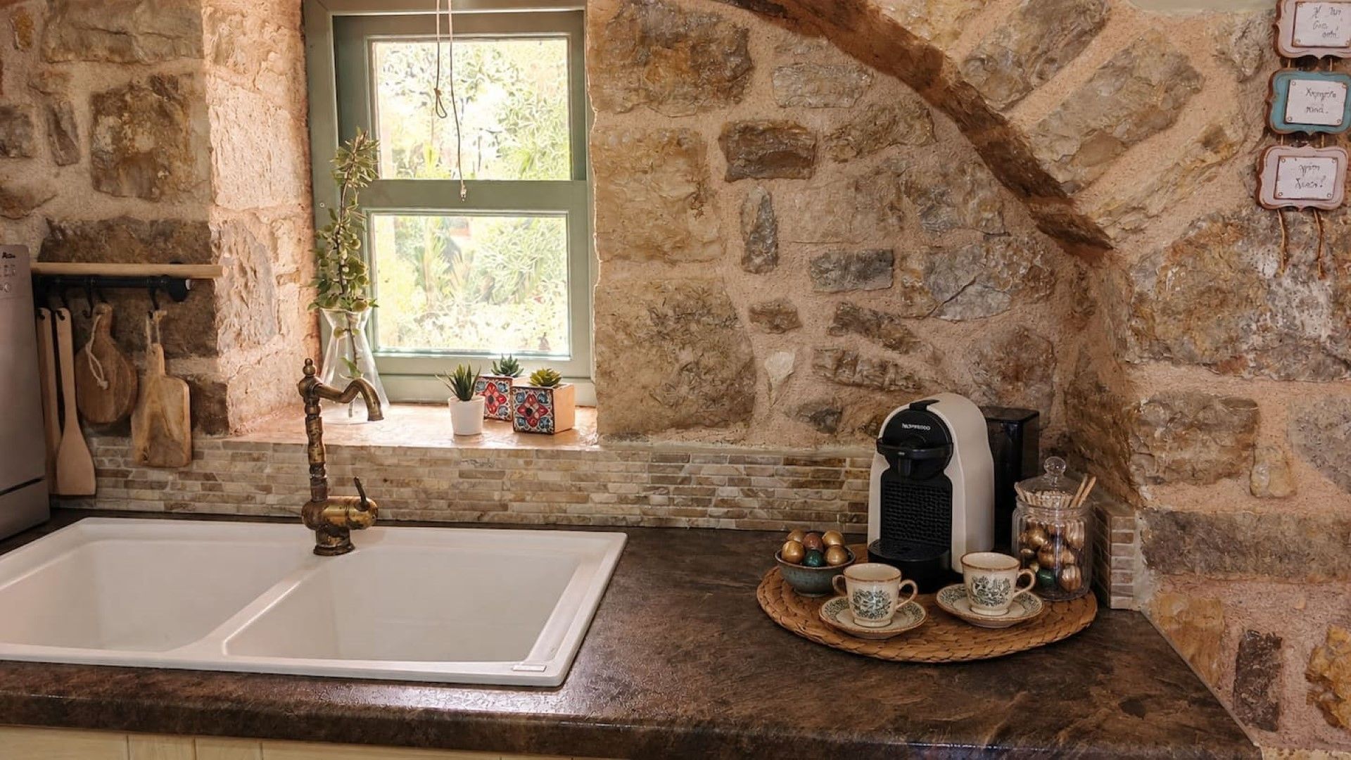 Uma cozinha com parede de pedra, janela com vegetação, máquina de café e pia.