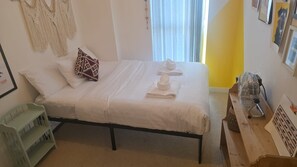 Una cama bien hecha con ropa blanca y una toalla doblada, un banco verde y una pieza de macramé colgada en la pared.
