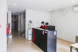 En reception med en person siddende bag disken, en vanddispenser, en brandslukker og en ventilator monteret på væggen.