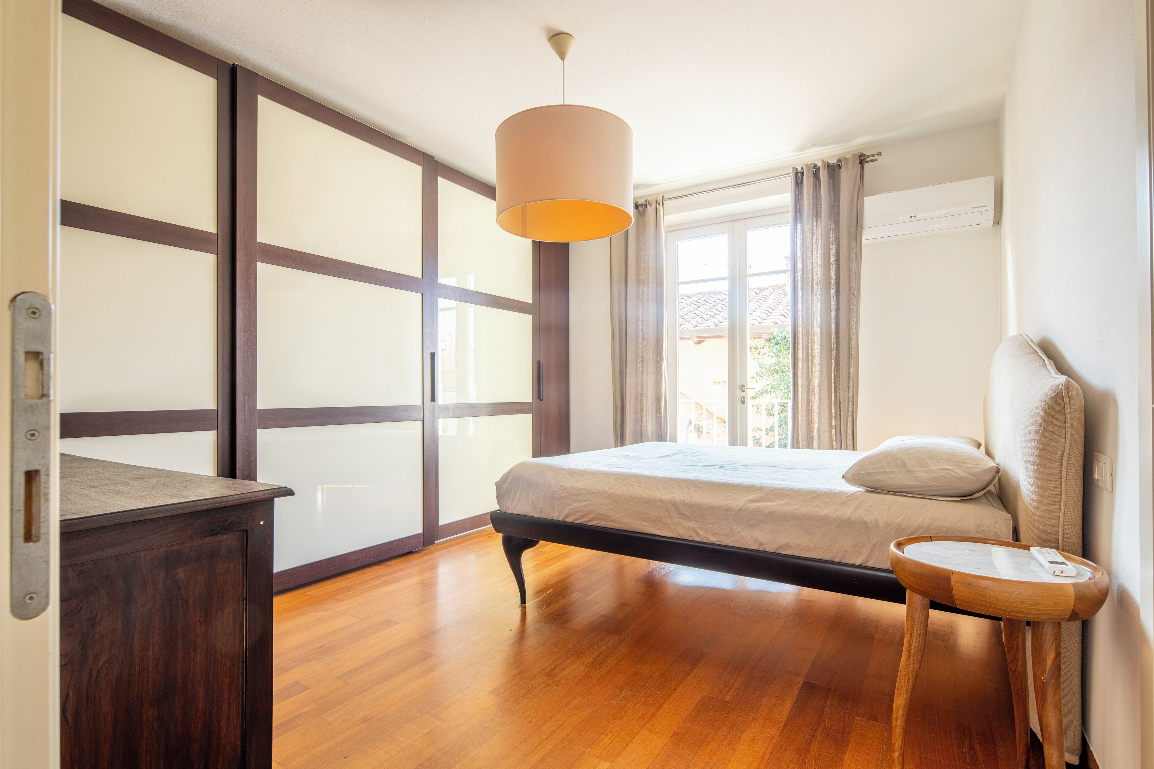 Un dormitorio con cama, mesita de noche, lámpara, armario y ventana con cortinas.