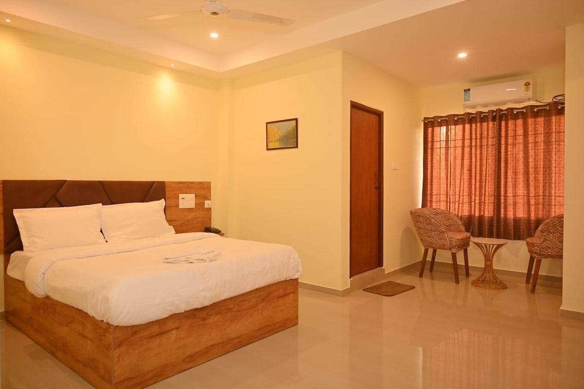 A hotel room with a bed, a seating area with two chairs and a table, a door, and an air conditioning unit.