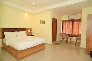 A hotel room with a bed, a desk, two chairs, a ceiling fan, and a door. - Vanveera Grand (Guwahati)