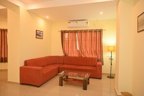 A living room with a curved sofa, a glass coffee table, a floor lamp, and a wall-mounted air conditioner. - Vanveera Grand (Guwahati)