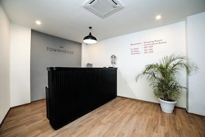 A reception area with a black reception desk, a potted plant, and a wall with room names and contact numbers. - Hotel O Elite Stayz (Gurgaon)