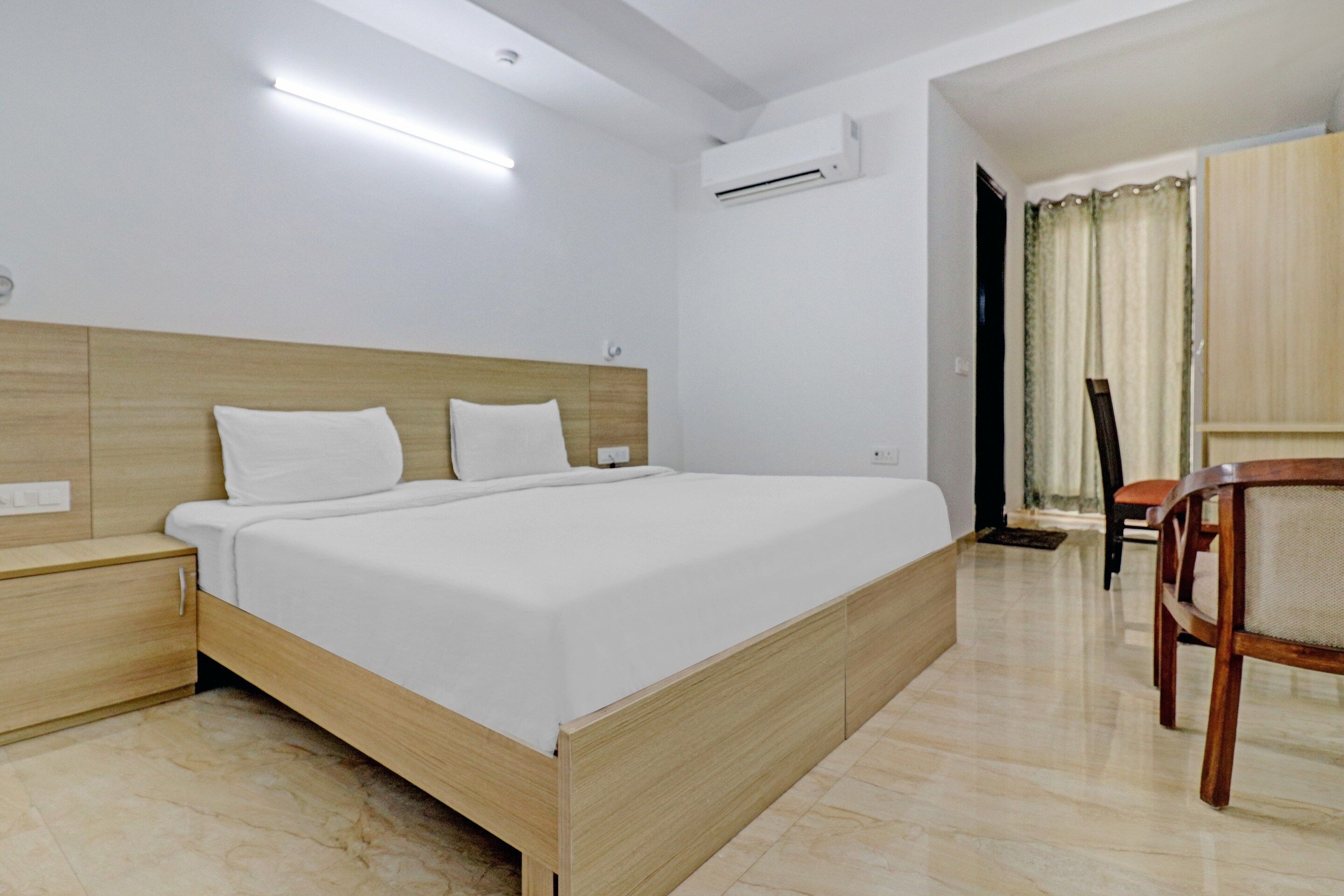 A hotel room with a wooden bed frame, white bedding, a nightstand, an air conditioning unit, and a wooden chair.