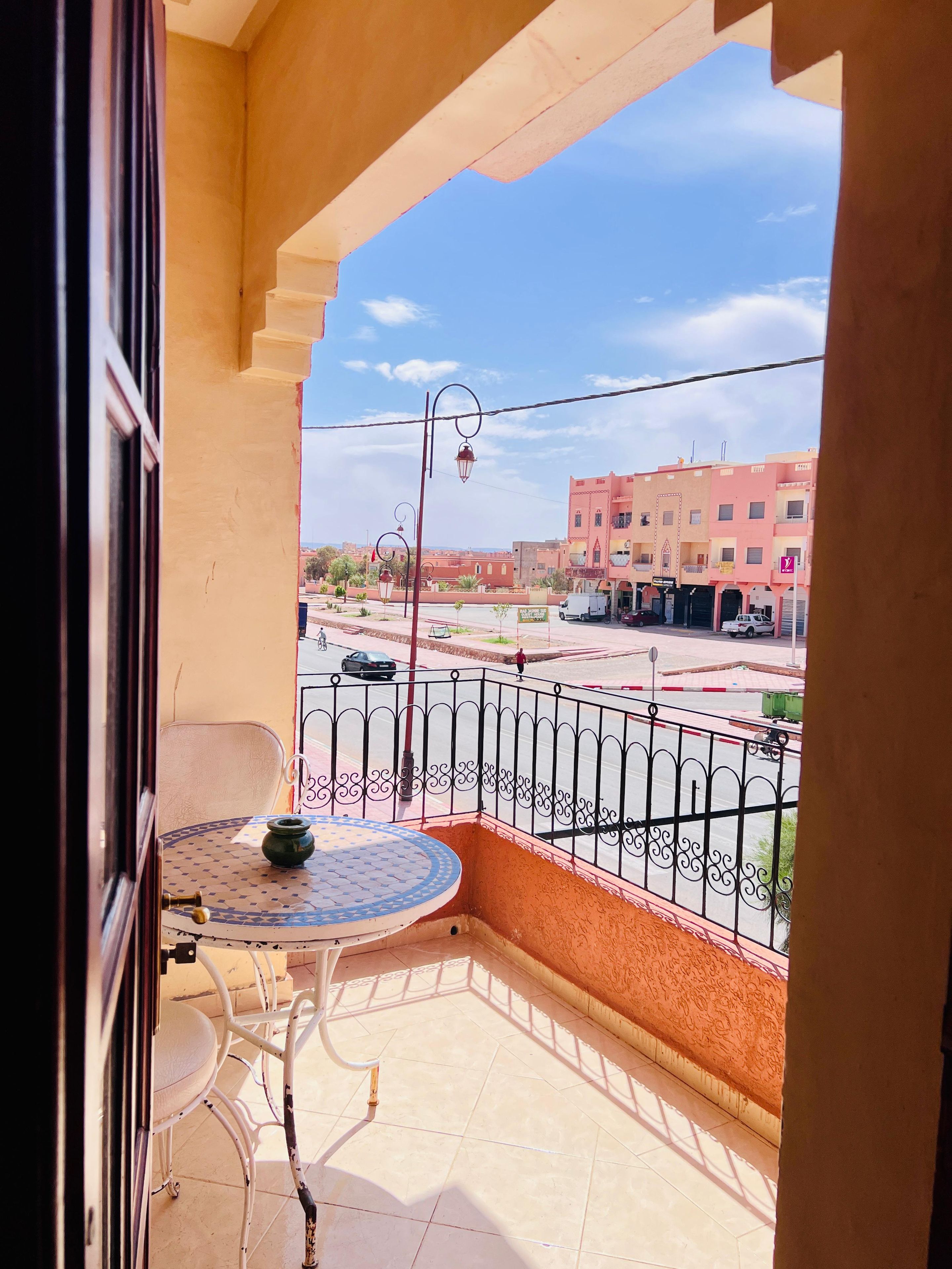 A balcony with a table and chairs, a view of a street with buildings, and a clear sky.