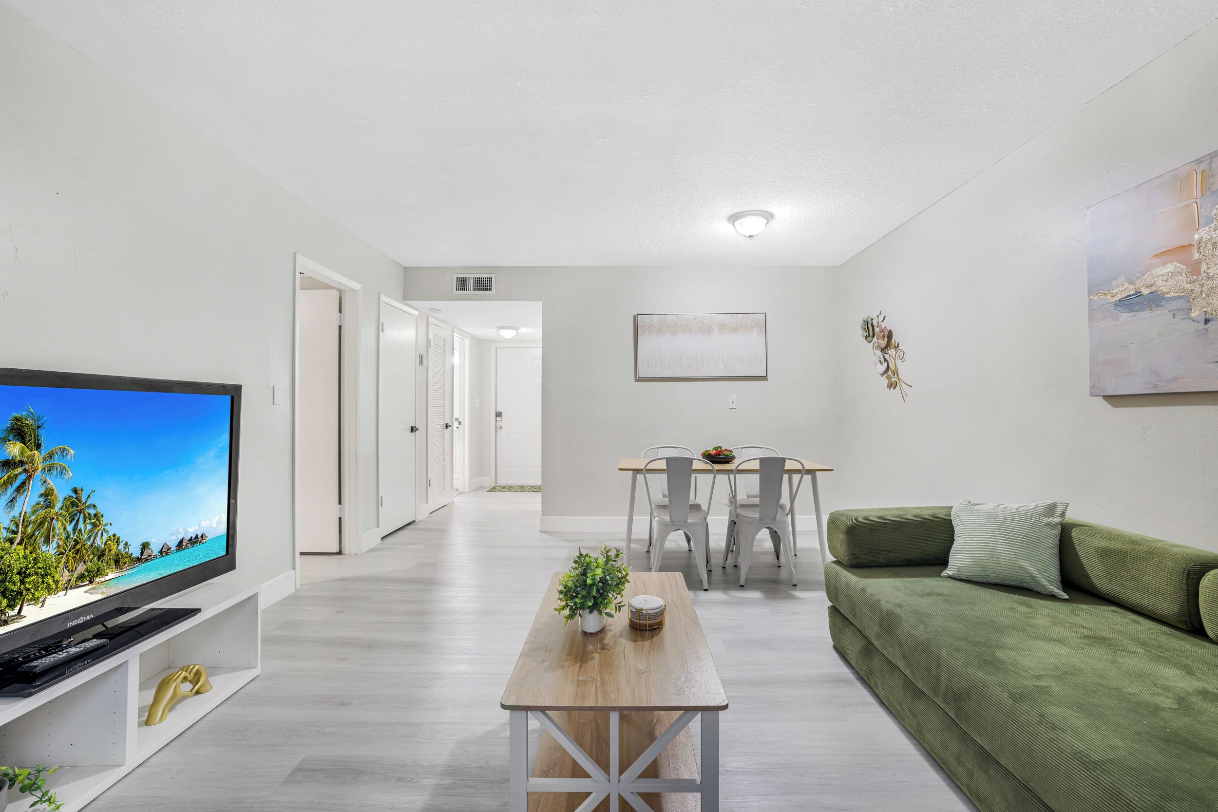 A modern living room with a flat-screen TV, a wooden coffee table, a green sofa, and a dining area with white chairs.