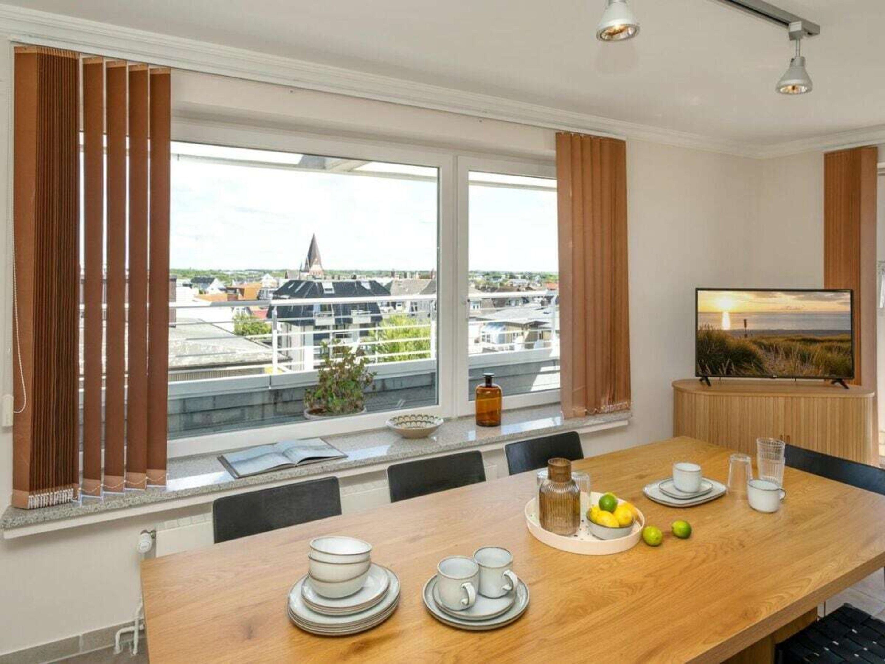 A dining area with a table set for two, a television on a stand, and a view of buildings through large windows.