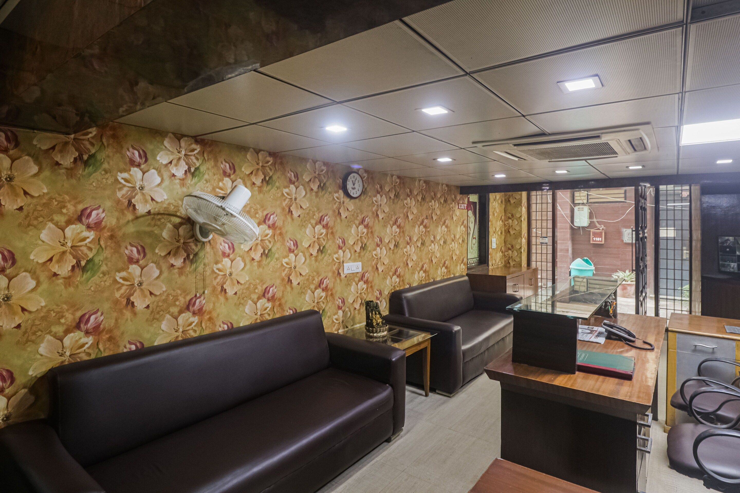Une salle d'attente avec un papier peint à motifs floraux, une horloge, un ventilateur, un canapé, un bureau et un téléphone.