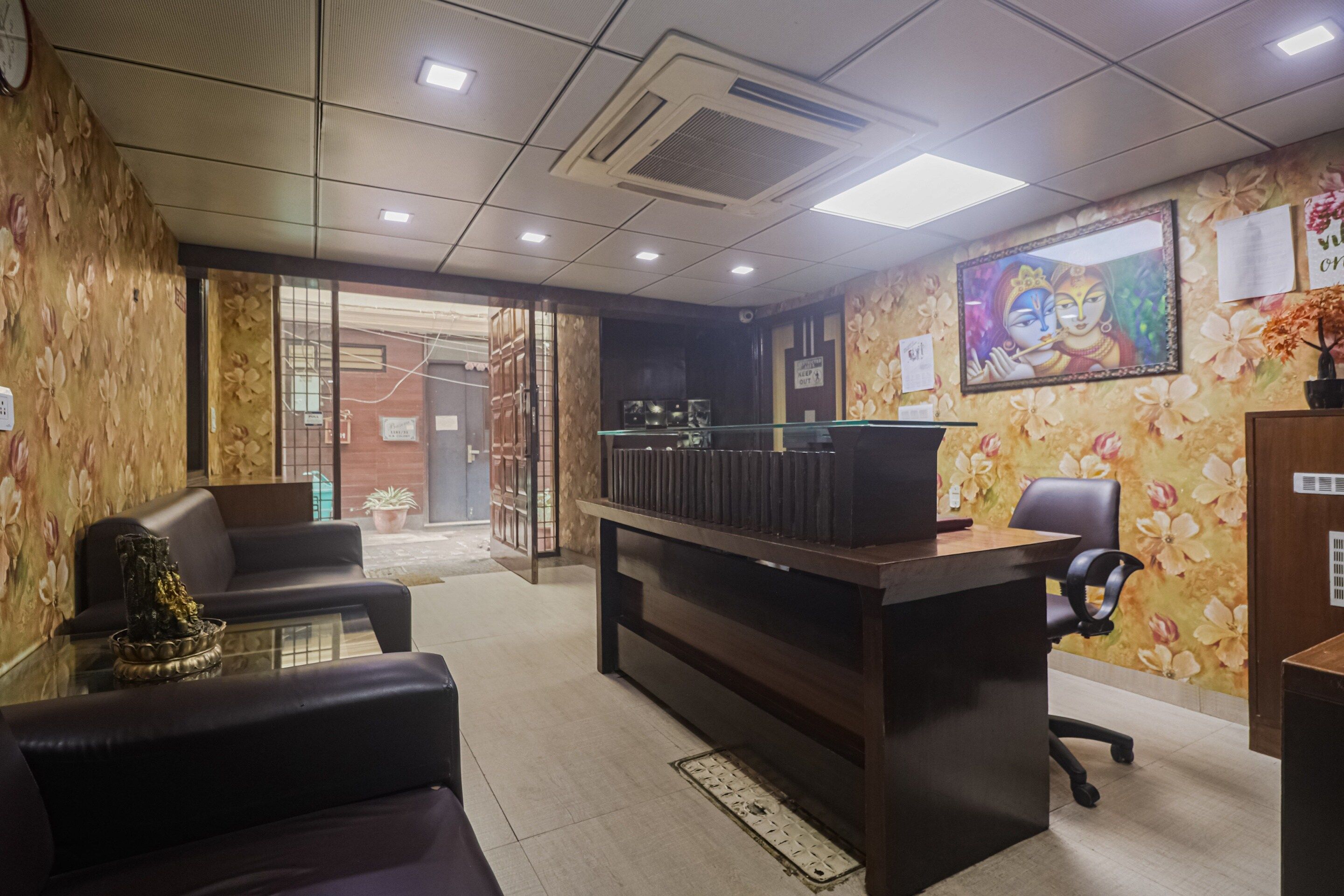 A reception area with a reception desk, a seating area with two chairs, a potted plant, and a wall with floral wallpaper and a framed picture.